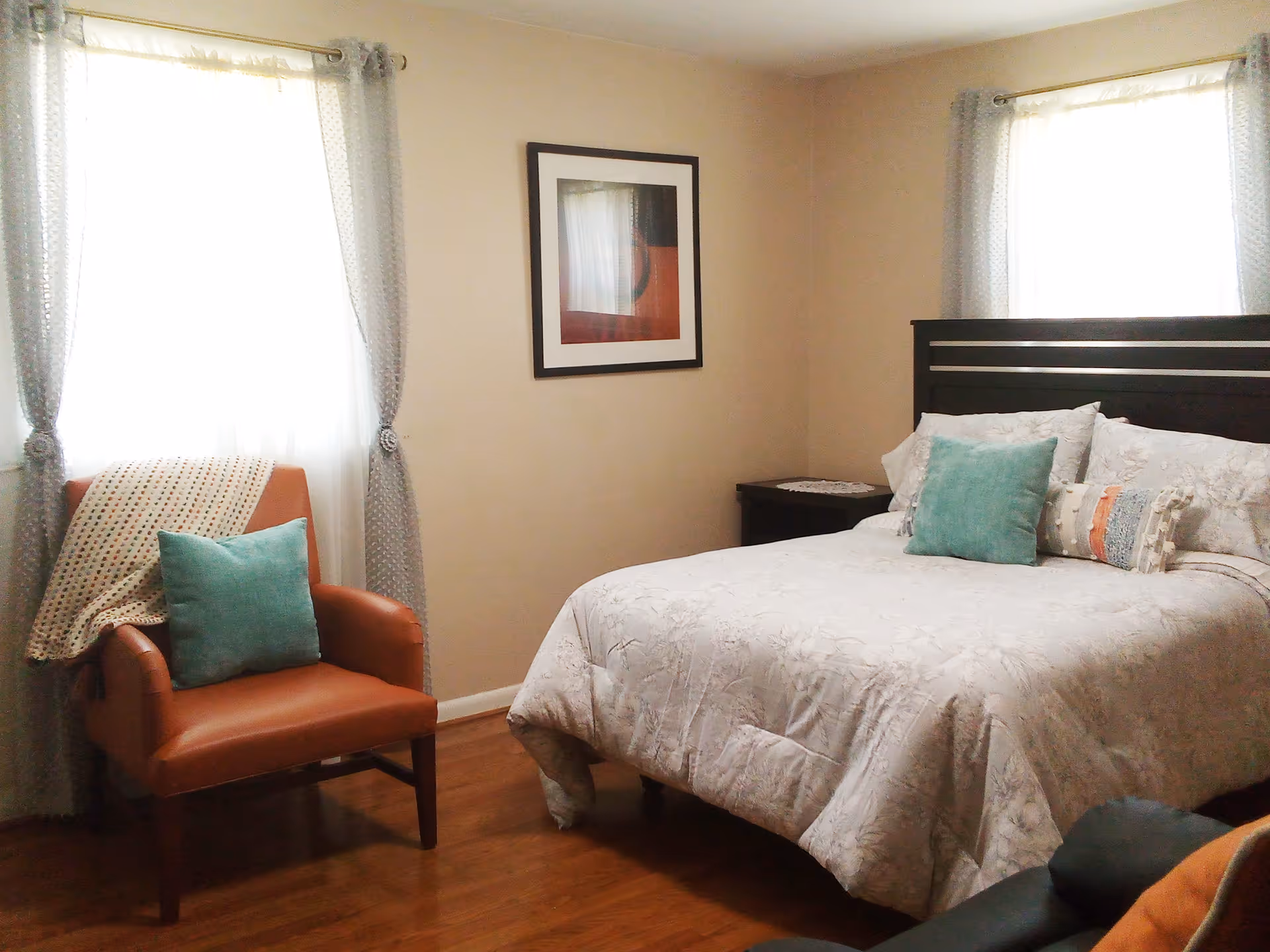 Bedroom with a large bed, teal pillows, chair by window, and framed wall art.