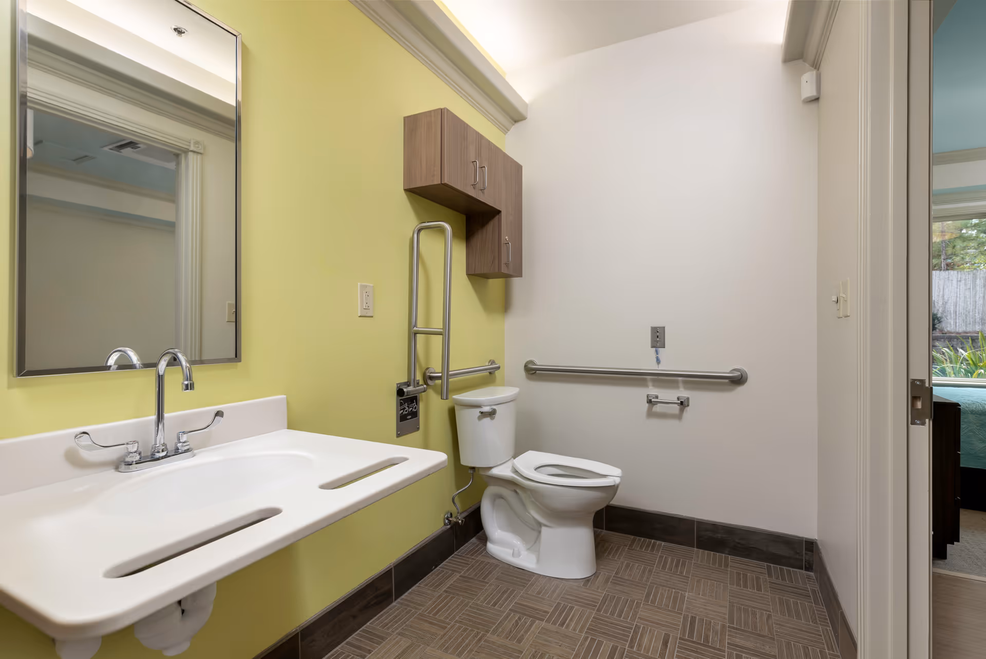 A clean, accessible bathroom with a white toilet and a wall-mounted sink with two elongated hand openings. The walls are painted light green and white, with a large mirror above the sink and a wooden cabinet mounted on the wall above the toilet. There are metal grab bars next to the toilet and on the adjacent wall for support. The floor has brown patterned tiles, and a doorway leads to a bedroom with a bed and window visible.