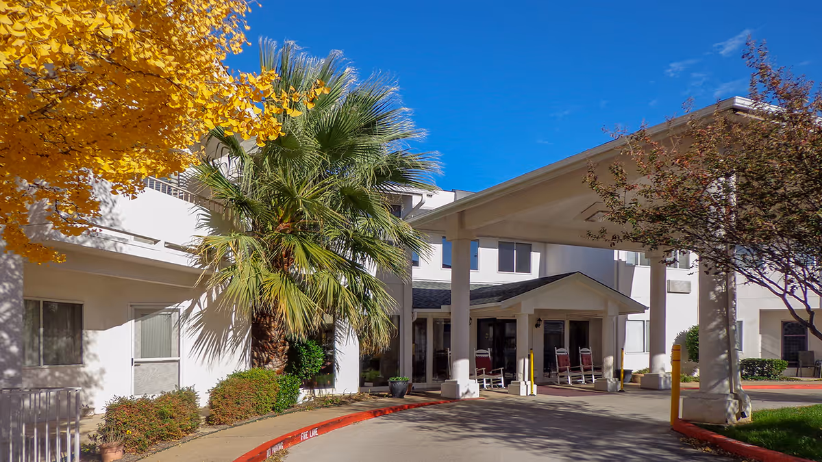 Entrance of Arlington Plaza - Sky Active Living facility with a covered drop-off area, palm trees, and other greenery under a clear blue sky.