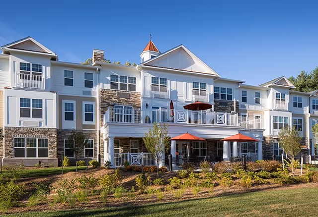 Exterior view of a large, multi-story senior living facility building with white and stone facade, multiple windows, balconies, and red umbrellas on the patio area. The building is surrounded by landscaped greenery and a clear blue sky.