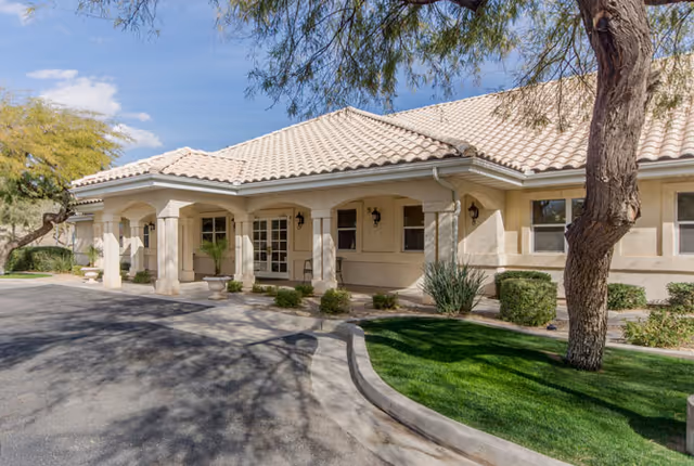 Single-story light-colored senior living facility entrance with a tiled roof, covered portico, and landscaped driveway.