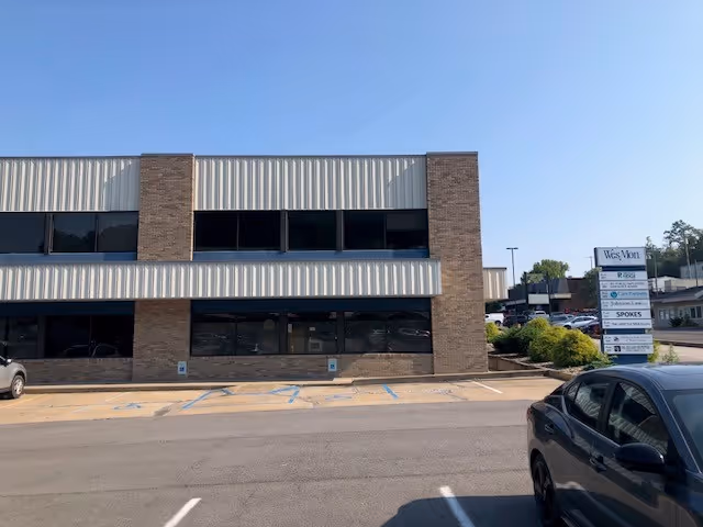 Two-story brick-and-metal commercial building with large windows and a parking lot in front.