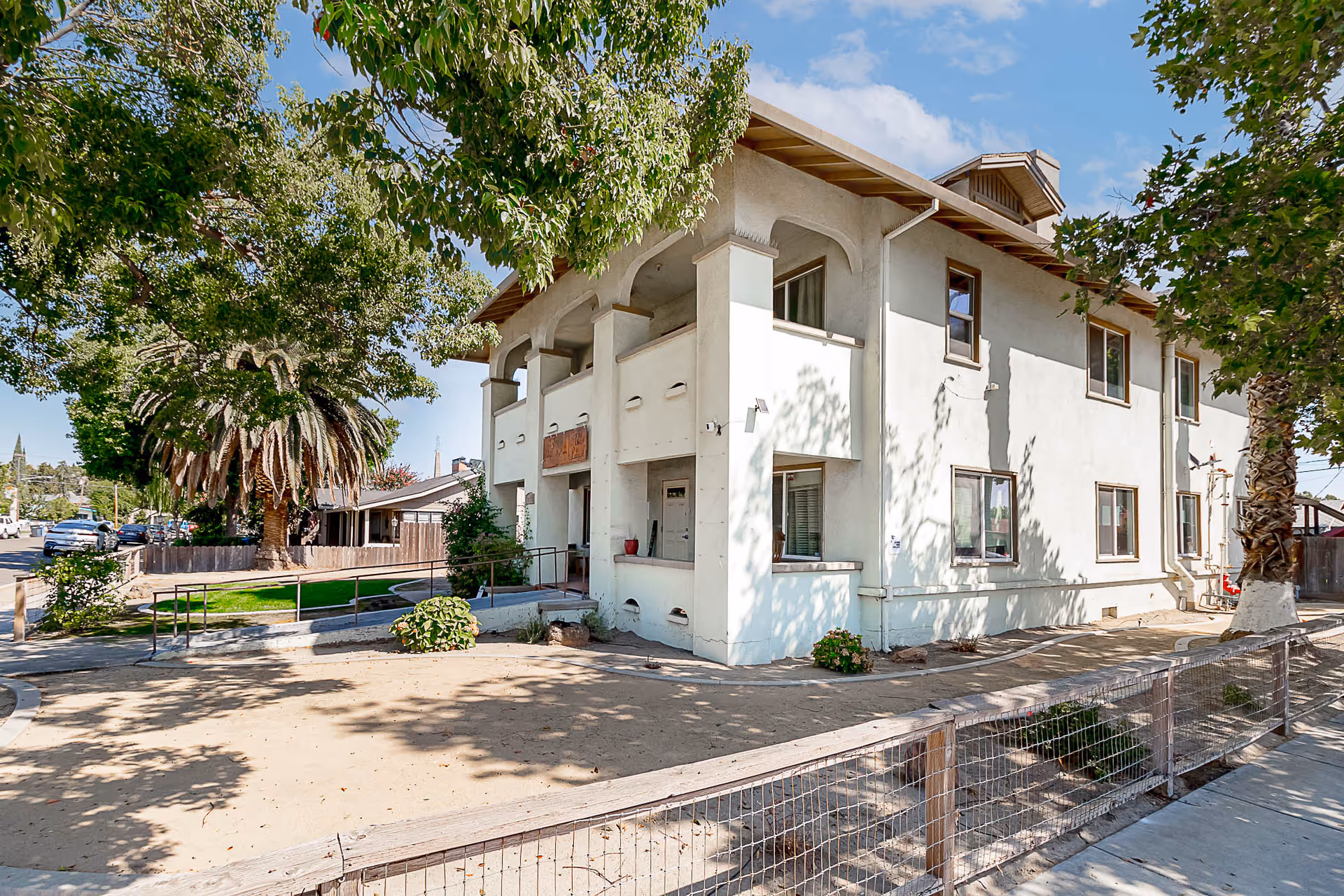 Two-story white senior living facility exterior with balconies, trees, and a sandy courtyard.