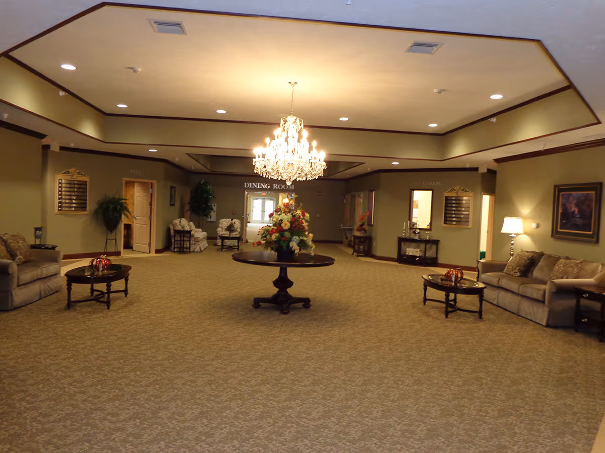 A spacious, well-lit common area with beige carpeting and light olive walls, featuring a large chandelier hanging from the ceiling. In the center, there is a round table with a floral arrangement. The room has several seating areas with sofas and armchairs, side tables with lamps, and decorative plants. A sign above a doorway in the back reads 'DINING ROOM'.