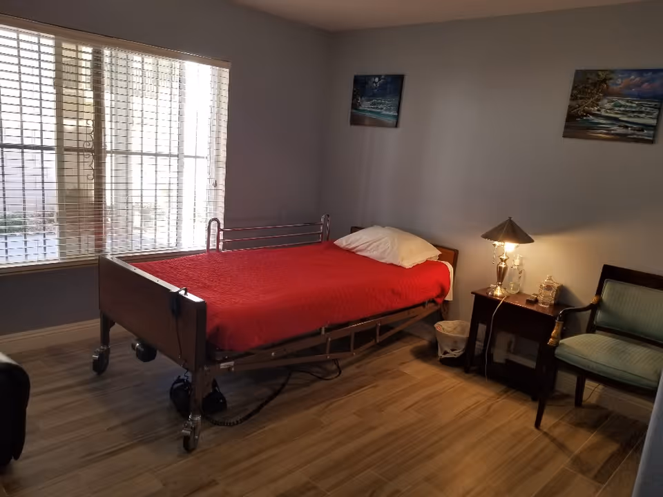 A hospital-style bed in a bedroom with red blanket, nightstand, lamp, chair, and window blinds