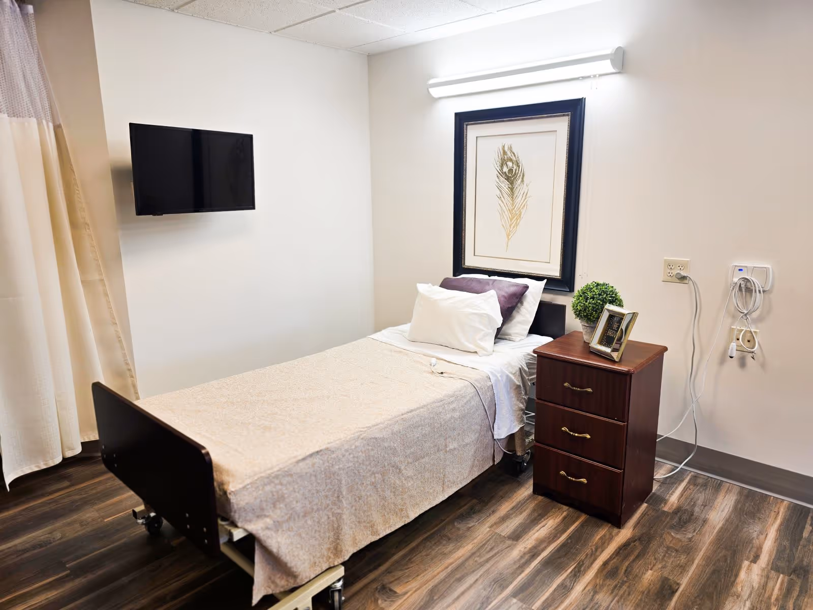 A furnished hospital-style bedroom with a single bed, dresser, wall art, and TV.