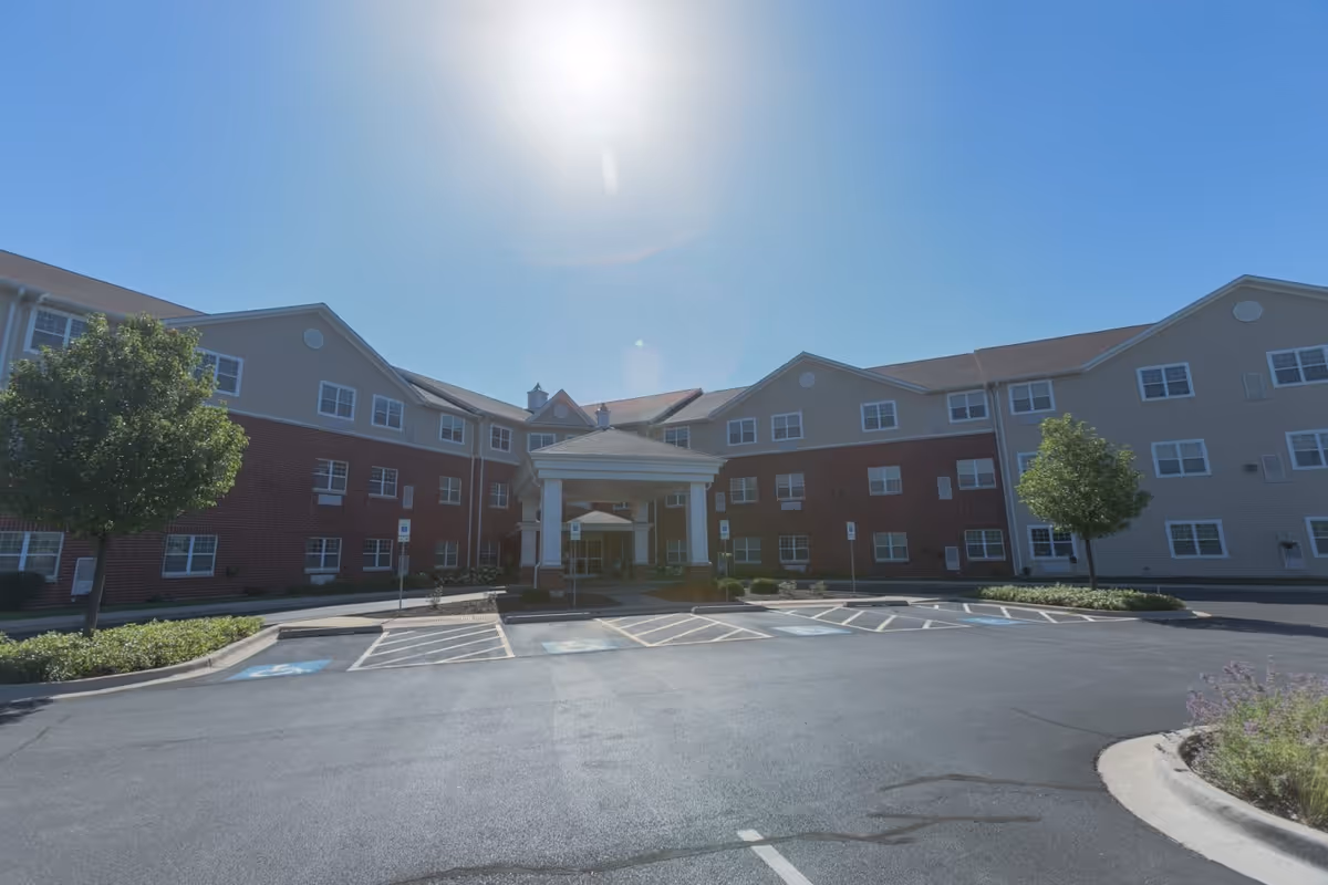 Exterior view of Heritage Woods of Plainfield, a multi-story senior living facility with a covered entrance, brick and beige siding, several windows, and a parking lot with handicap spaces in front. The sky is clear and sunny.