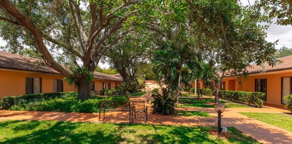 Sunlit courtyard with benches, large trees and walkways flanked by single-story peach-colored residential buildings.