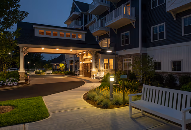 Evening view of the entrance to a senior living facility with a covered driveway, illuminated pathway lights, benches, and a multi-story building with balconies.