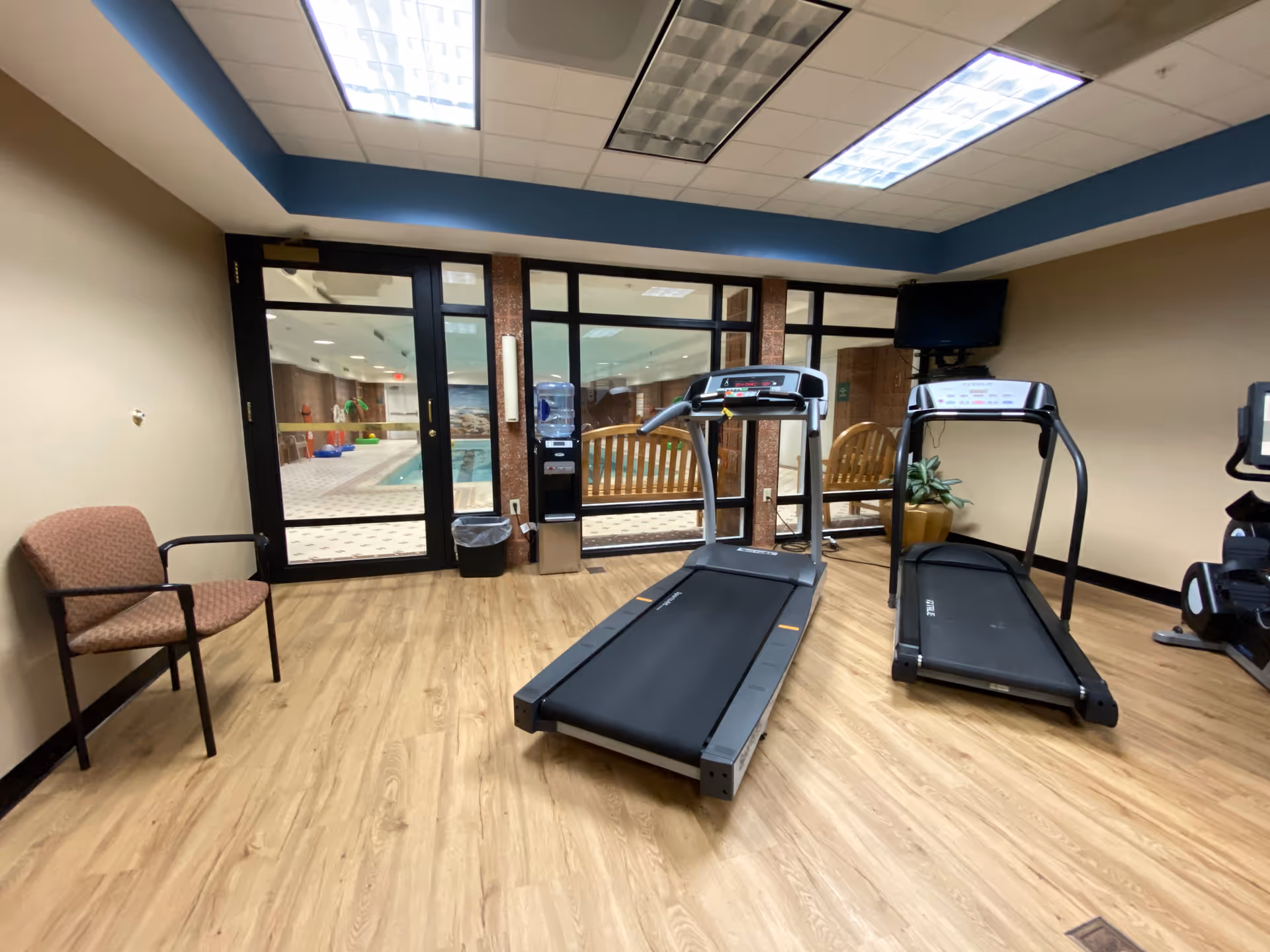 Interior fitness room with treadmills, chairs, and a view of the pool area through glass doors.