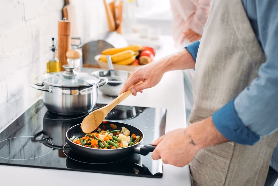 A person wearing an apron is cooking mixed vegetables in a frying pan on a stovetop in a bright kitchen. There is a pot with a lid on another burner, and a bowl of bananas and other fruits on the counter in the background.