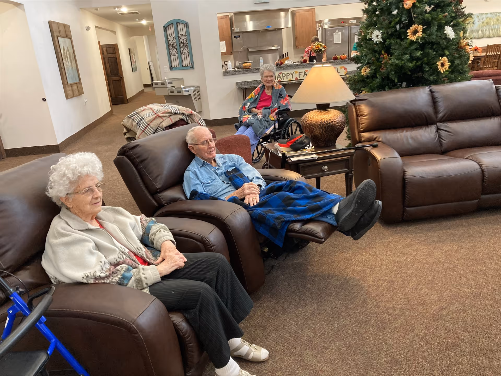 Three elderly individuals sitting in a common living area of a senior living facility. Two are seated in brown leather recliners, while one is in a wheelchair. There is a large decorated Christmas tree, a table with a lamp, and a kitchen area in the background with a 'Happy Fall' banner. The room has carpeted floors and neutral-colored walls.