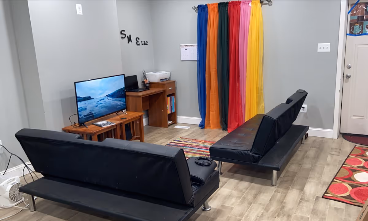 A small living room area with black couches, rainbow curtains, and a TV.