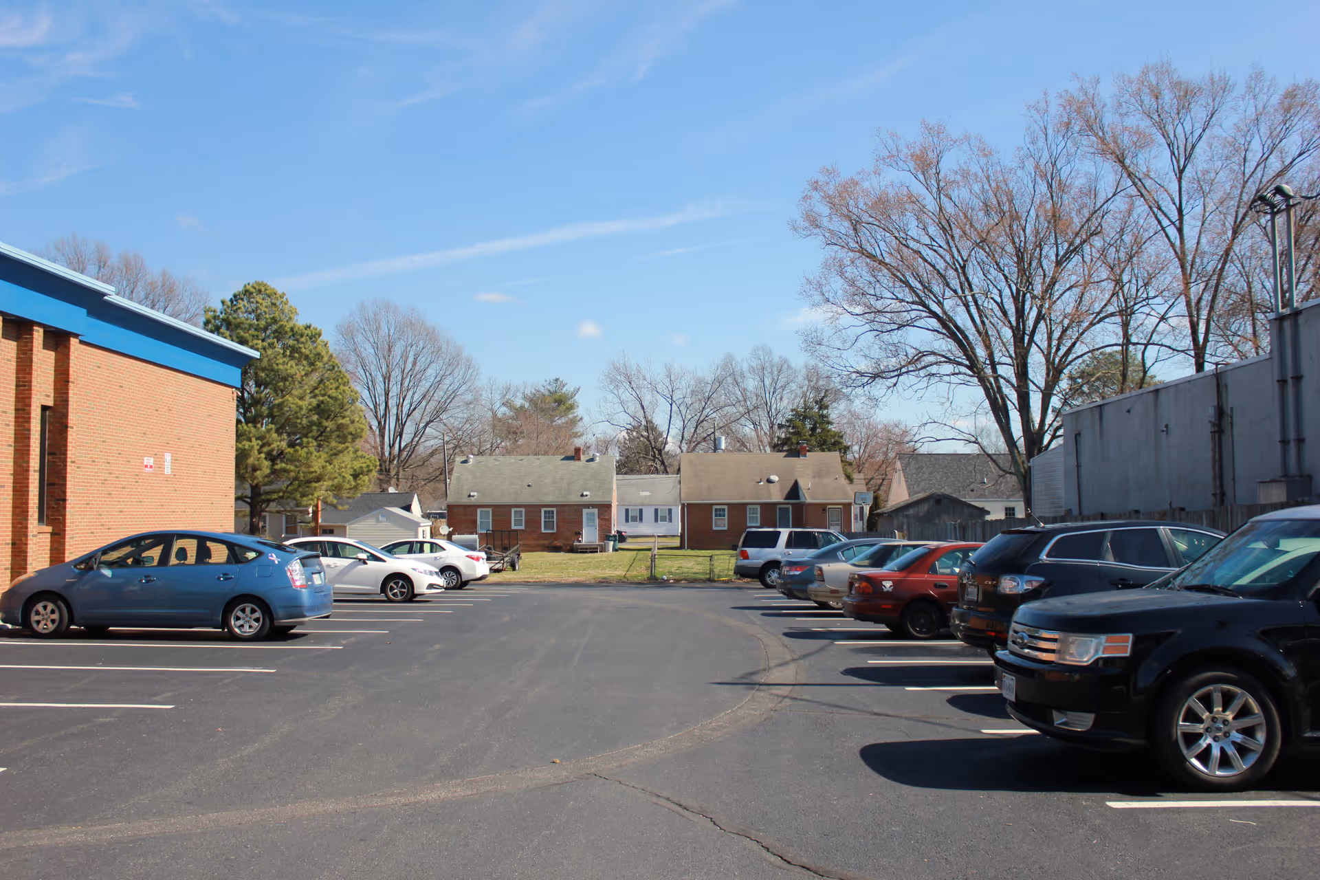 Exterior parking lot with cars, a brick building, and leafless trees under blue sky.