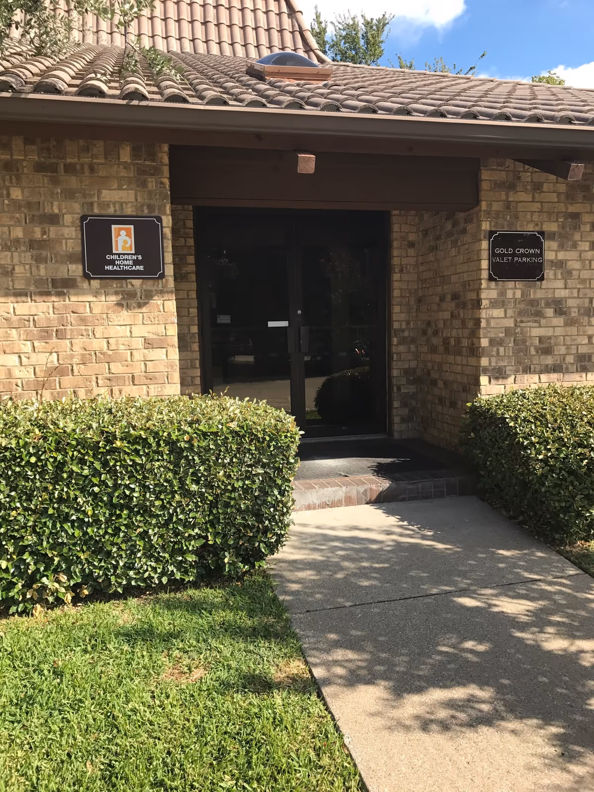 Brick facility entrance with glass doors, signage, and trimmed hedges on a sunny day.