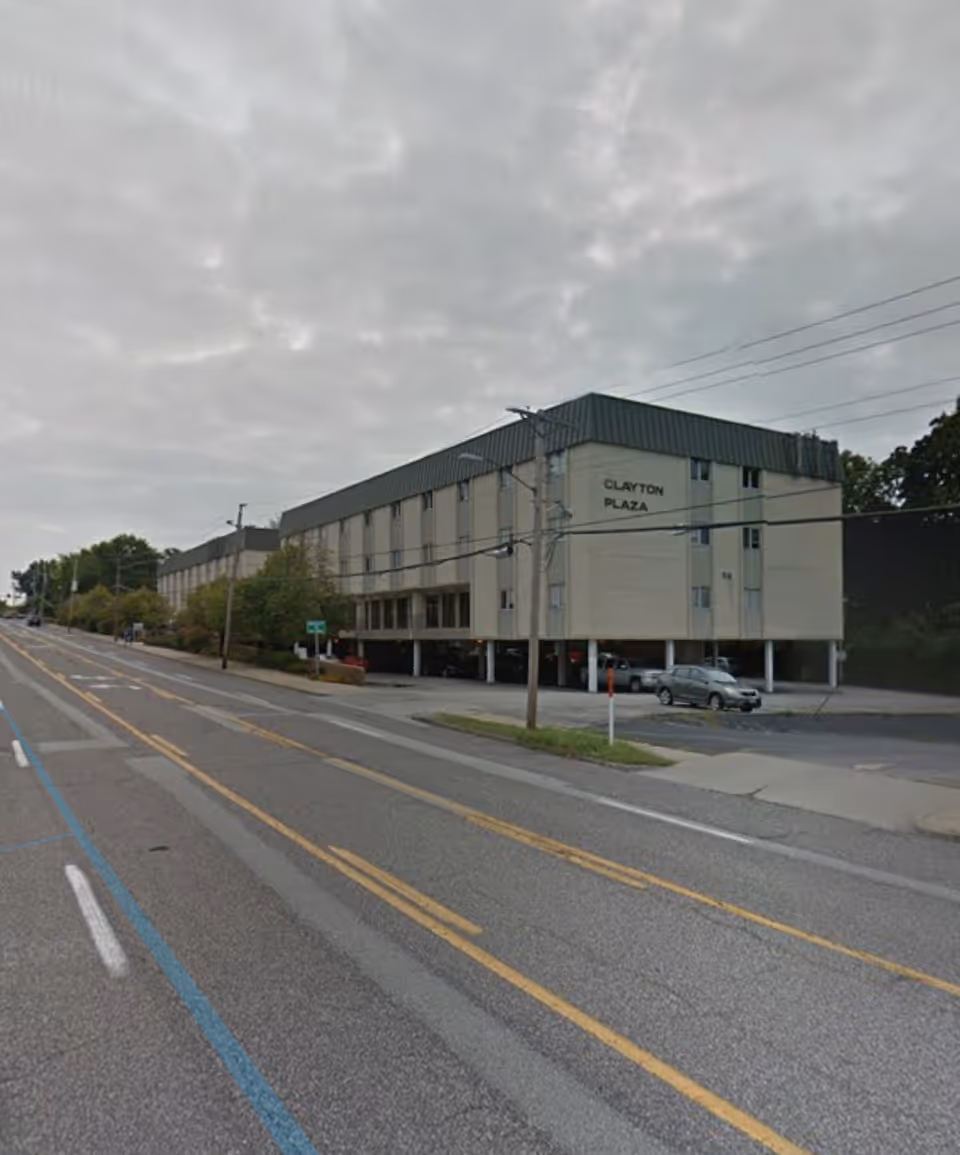 Exterior view of a beige, multi-story building labeled Clayton Plaza beside a street with parked cars.