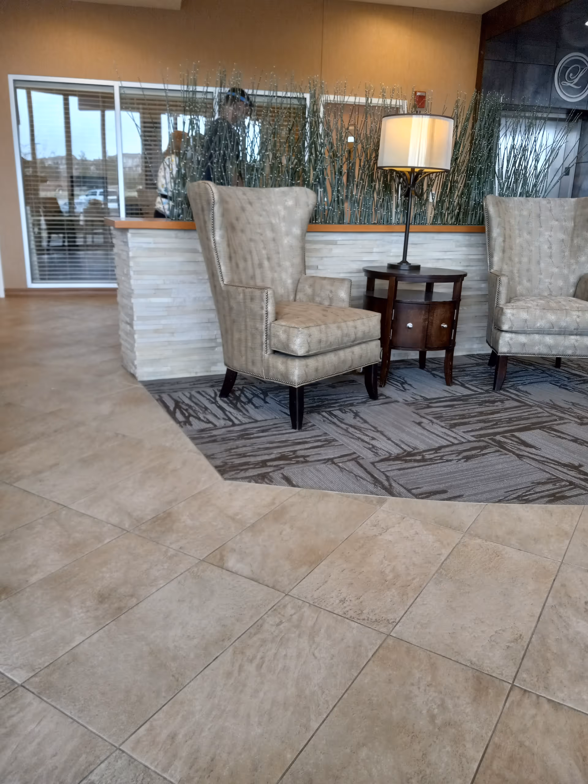 Two beige high back chairs flank a round wooden table with a lamp in a lobby.