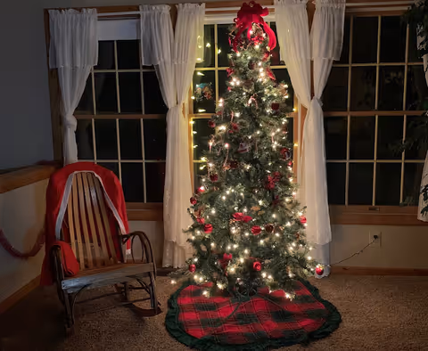 A decorated Christmas tree with white lights, red ornaments, and a red bow on top stands in front of a window with white curtains. To the left of the tree is a wooden rocking chair draped with a red and white Santa Claus coat. The room has beige carpet and wooden trim around the windows and walls.