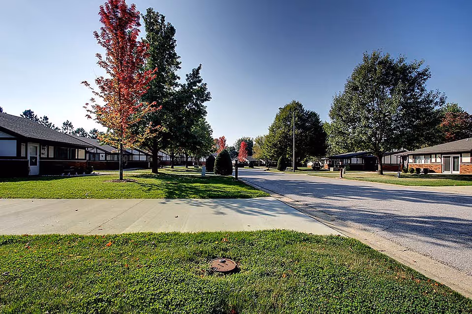 Street view of a single-story senior living complex with lawns, trees, sidewalks, and a clear sky.