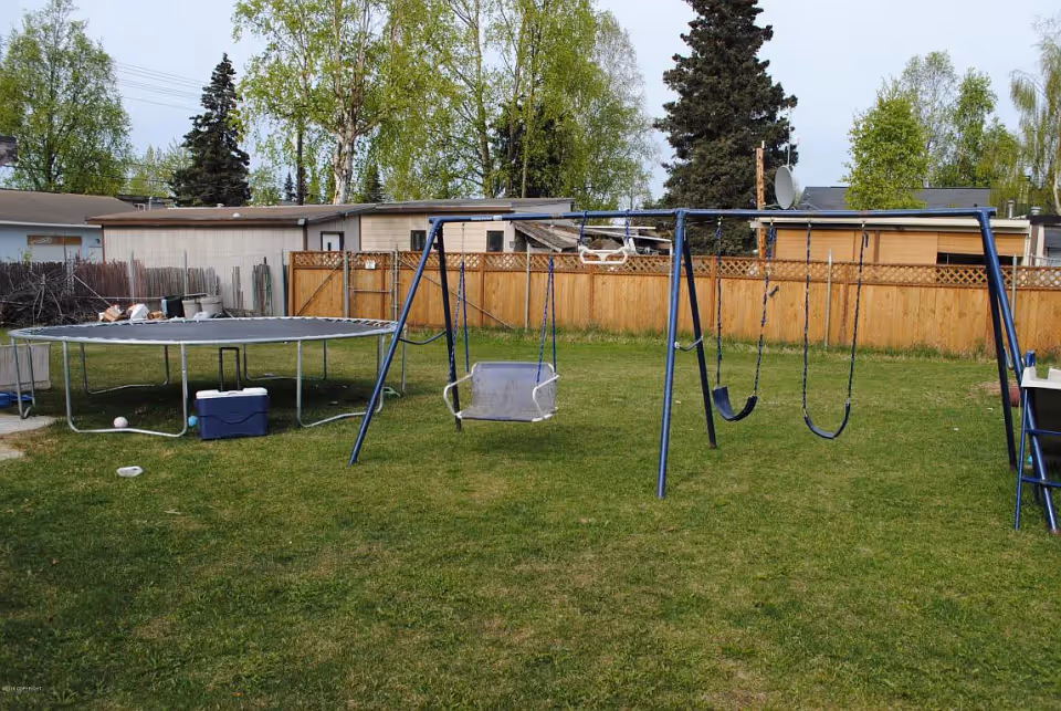 A backyard with a green lawn featuring a trampoline on the left and a blue swing set with multiple swings in the center. There is a wooden fence and some trees in the background, along with small buildings or sheds.