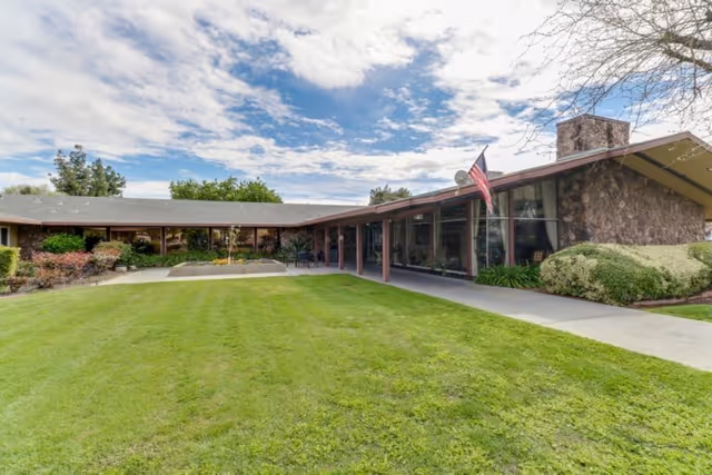 Single-story care facility front with a wide green lawn, covered walkway, large windows, and an American flag under a partly cloudy sky.