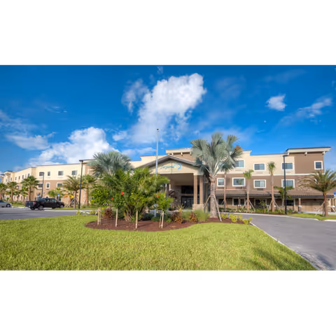 Front exterior of a three-story senior living building with a covered entrance, landscaped lawn and palm trees under a blue sky.