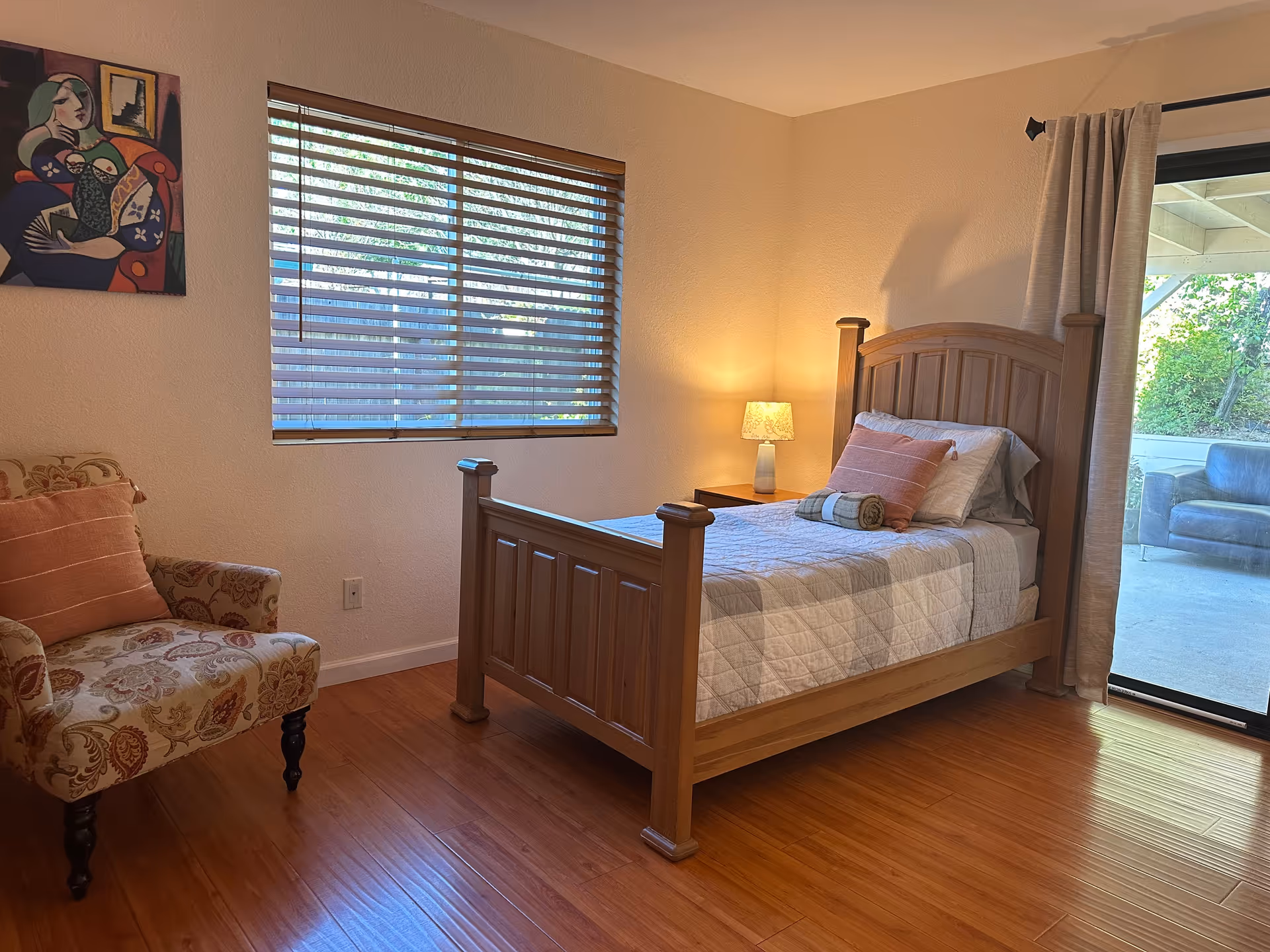 Cozy bedroom with wooden bed, chair, nightstand lamp, blinds, and sliding glass door.