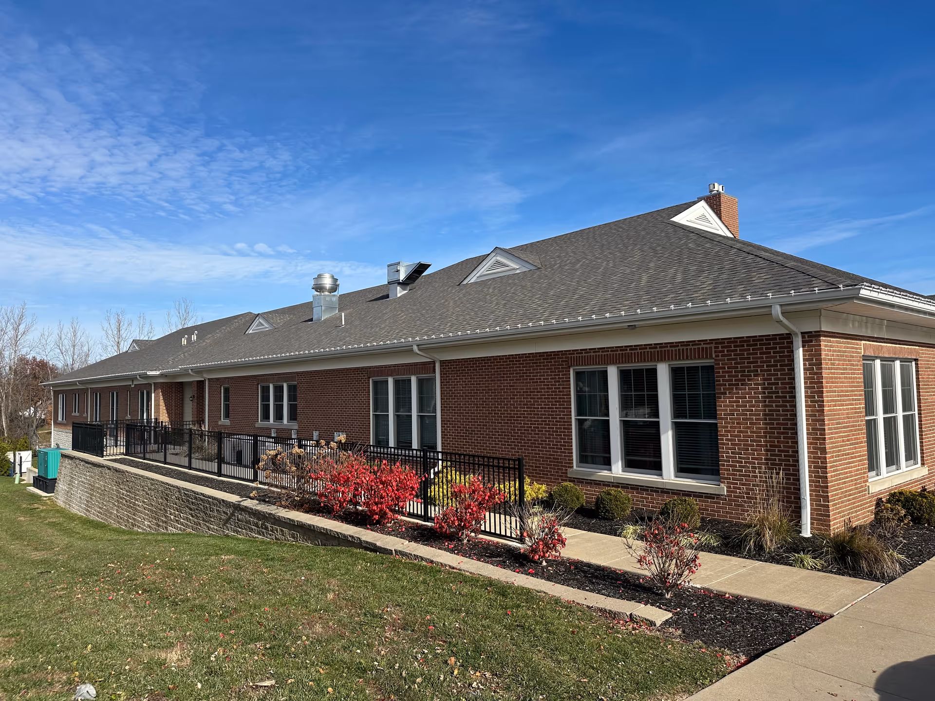 Single-story brick senior living building exterior with windows, a ramped walkway, railing and landscaped flower beds under a blue sky.