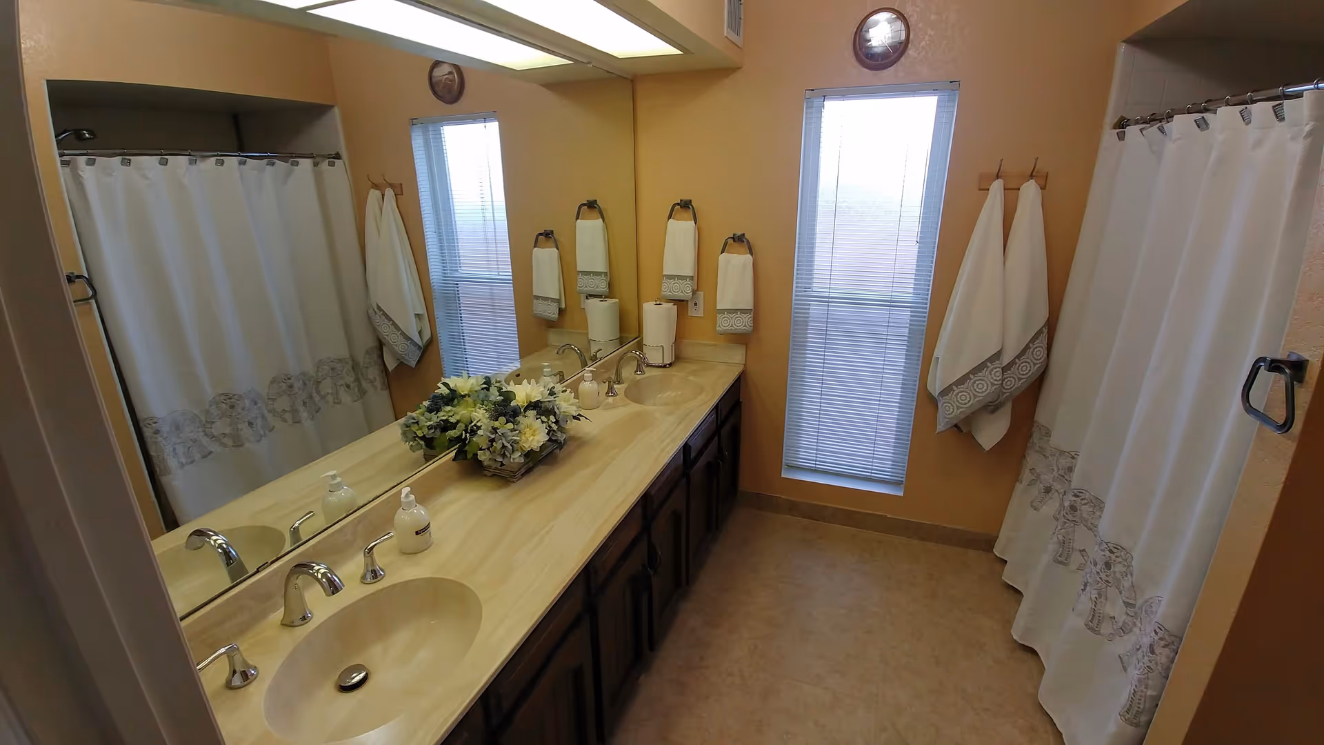 Bathroom in a senior living facility with double sinks, mirrors, towels, and a shower curtain.