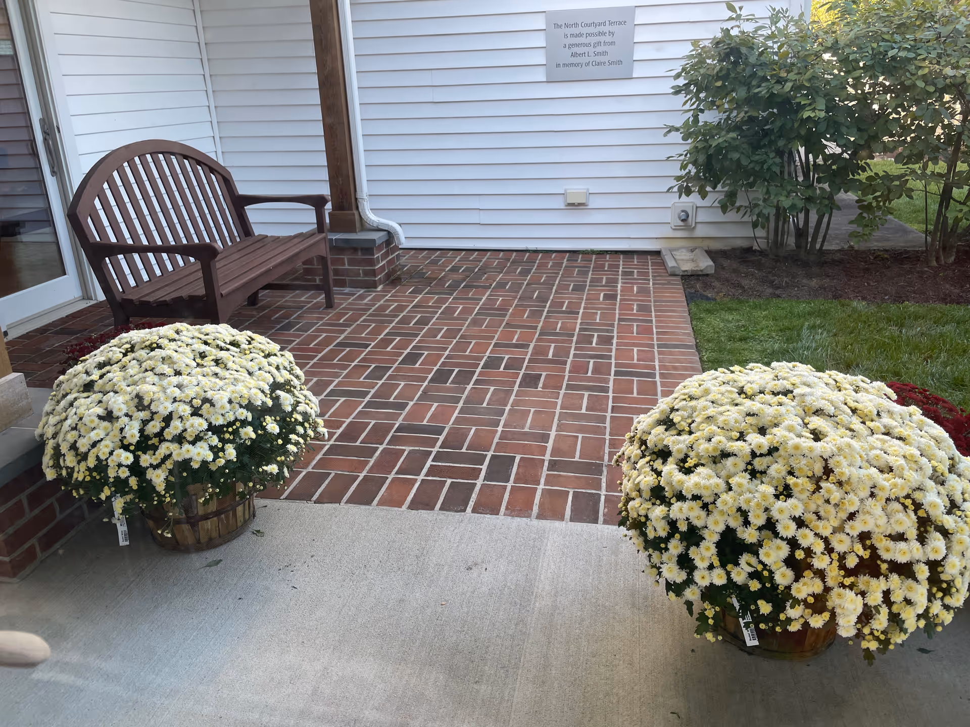 Brick patio with bench, potted white chrysanthemums, and a house exterior.