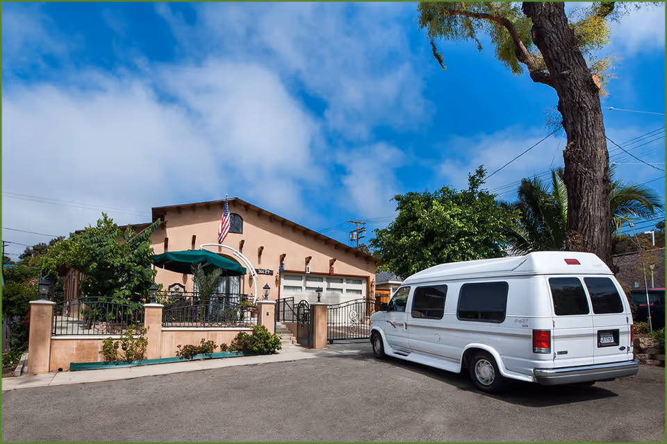 Front exterior of a stucco senior living building with a gated entrance, an American flag, and a white van parked in front.