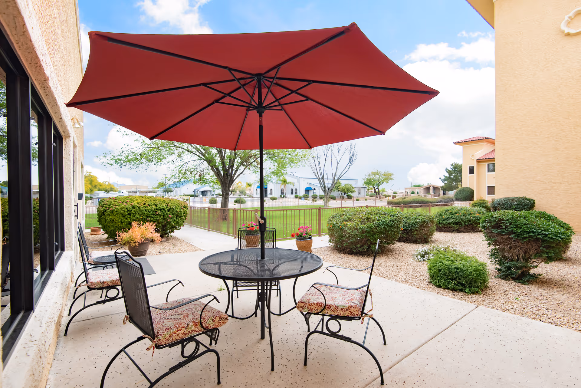 Patio with a round metal table, four chairs and a large red umbrella overlooking a landscaped courtyard and lawn.