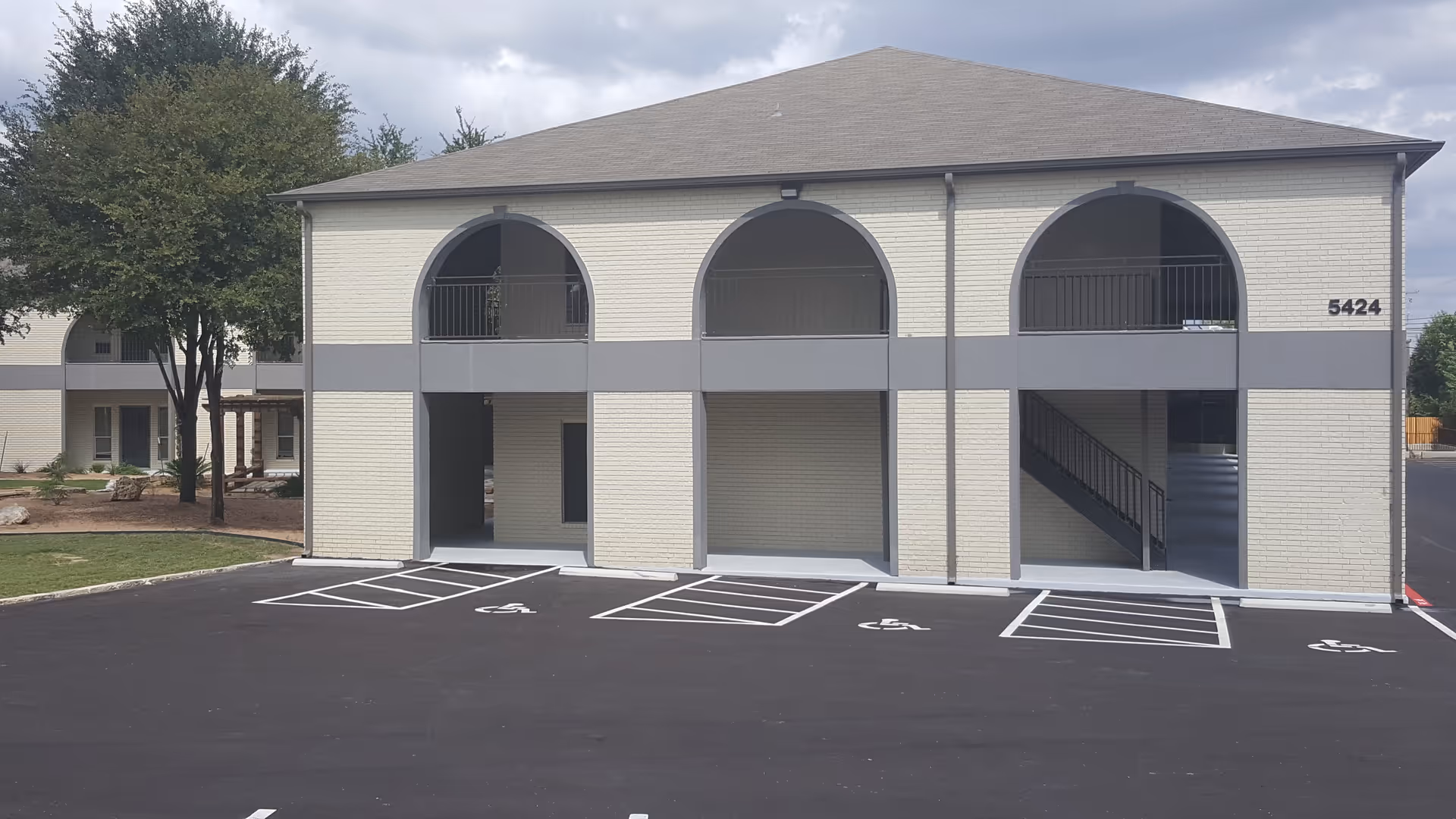 Two-story light-brick senior living building with arched openings and a front parking lot.