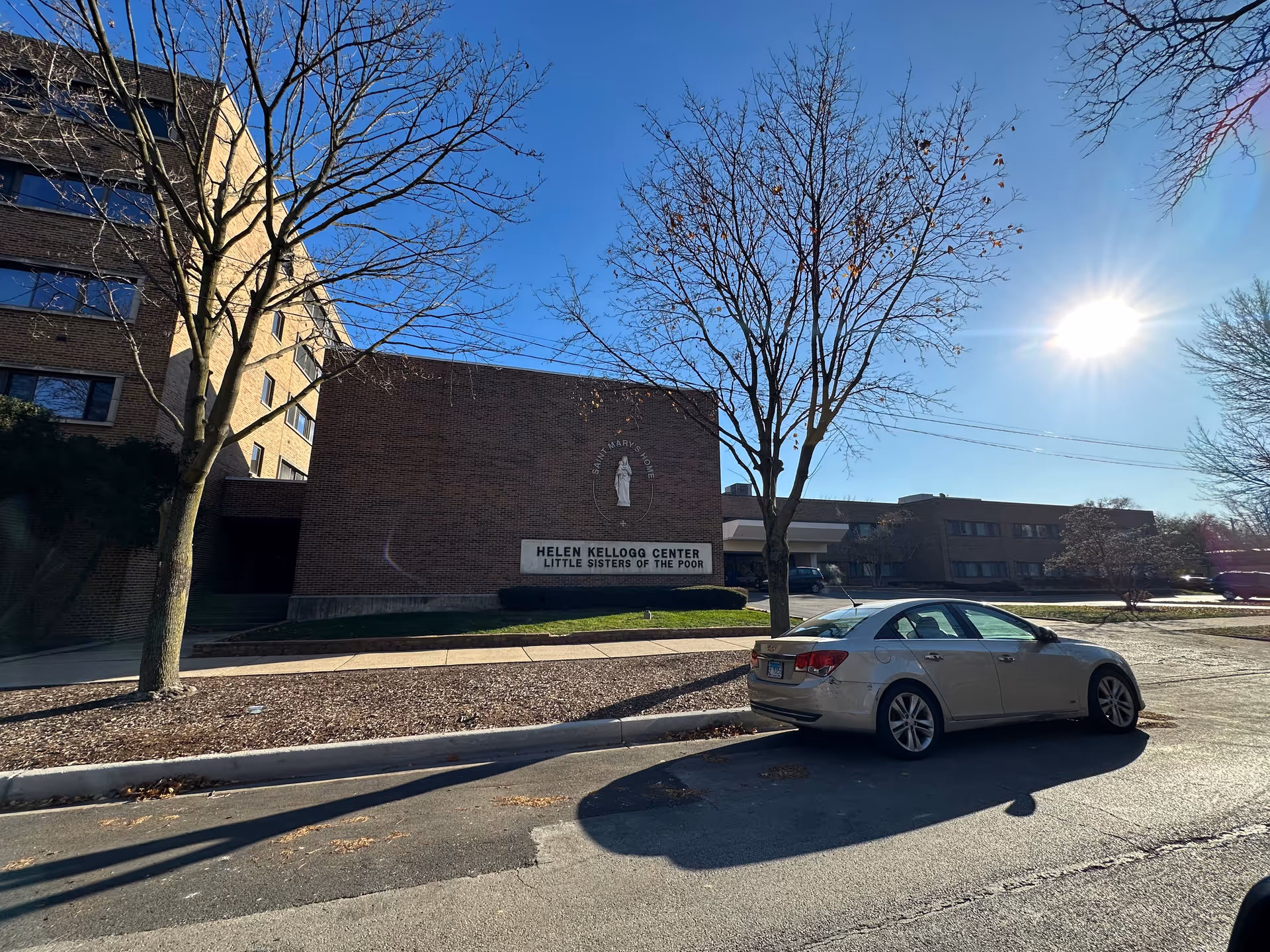 Front exterior of a brick senior living building with a sign, leafless trees, a parked car, and the bright sun in a clear blue sky.