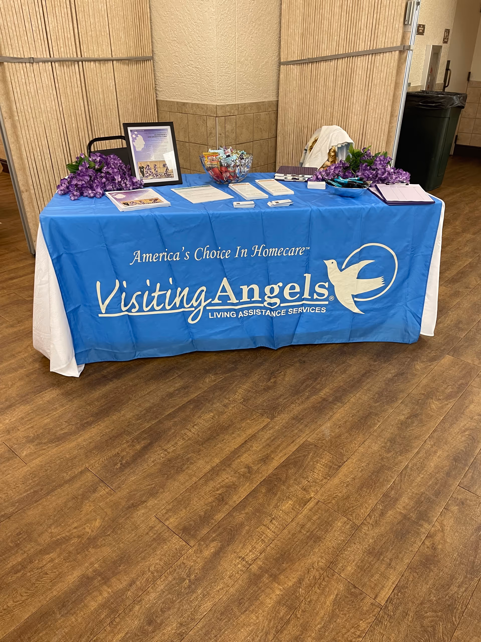 Blue-covered table with Visiting Angels banner stands in an indoor corridor.