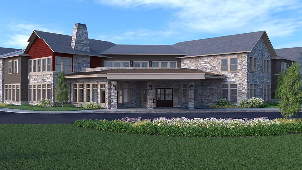 Exterior view of a two-story assisted living facility building with stone and wood siding, a covered entrance, and landscaped greenery including bushes and flowers in front.