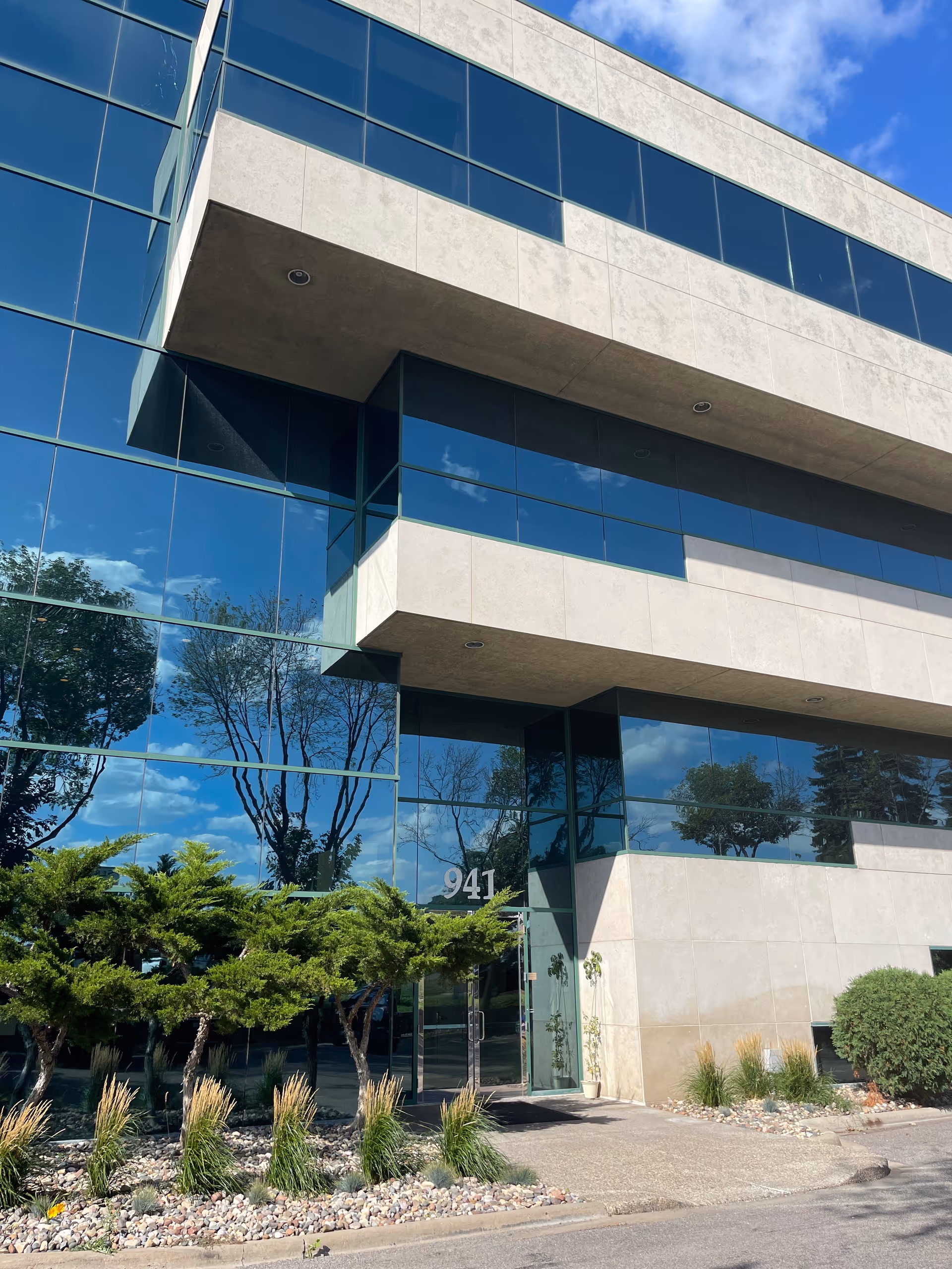 Modern glass-front office building exterior with landscaped entrance and greenery.