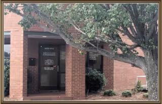 Brick building entrance with glass doors, tree, and shrubs in front.