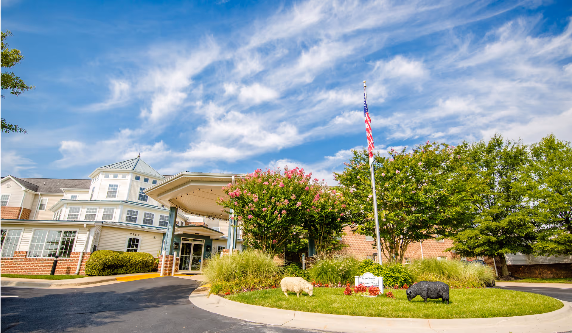 Exterior view of Arbor Terrace Sudley Manor with entrance canopy, landscaping, and American flag