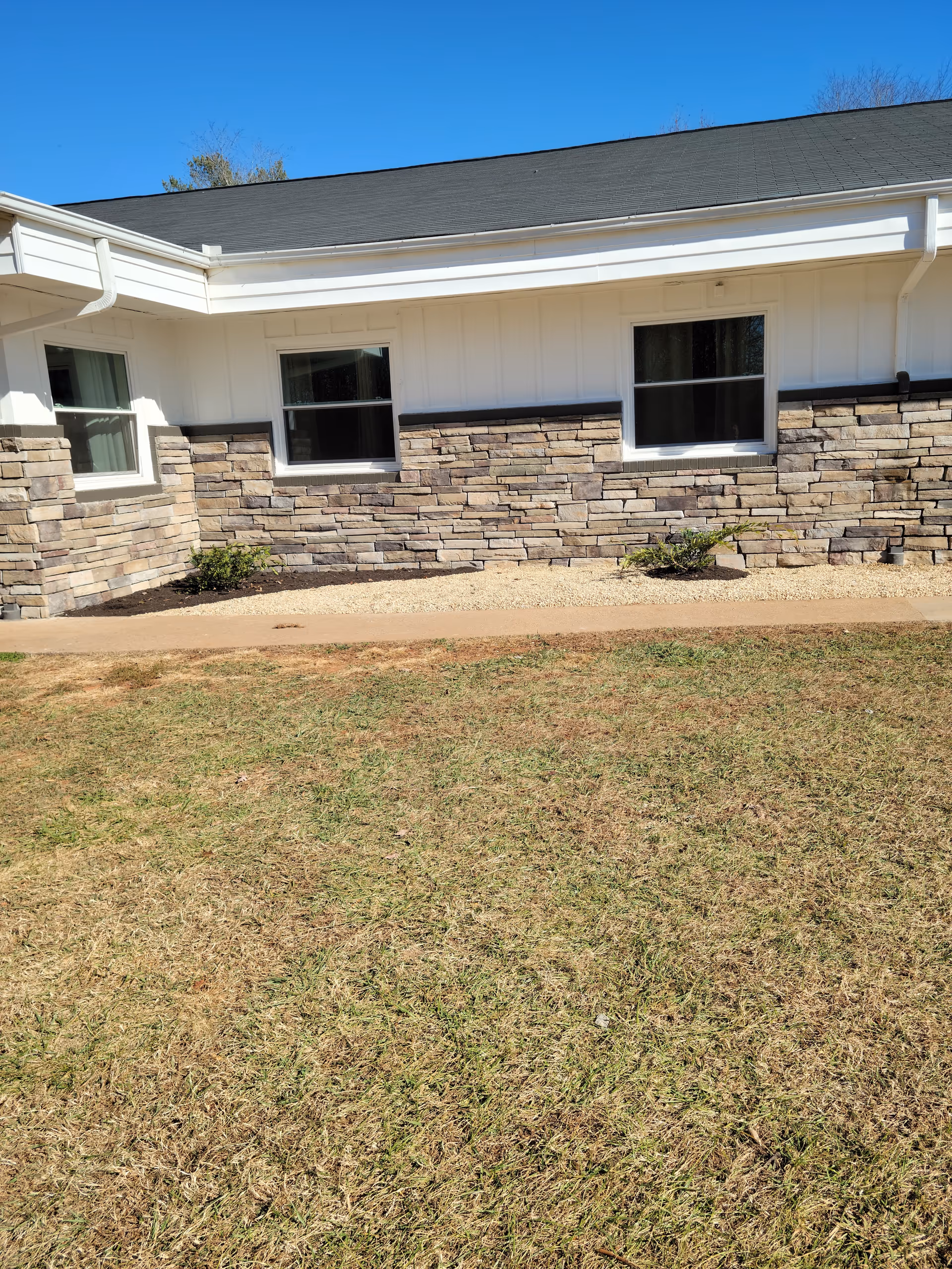 Exterior view of a single-story building with white siding and stone accents below the windows. There are three windows visible, a small garden bed with mulch and small shrubs, a concrete walkway, and a grassy lawn in the foreground under a clear blue sky.