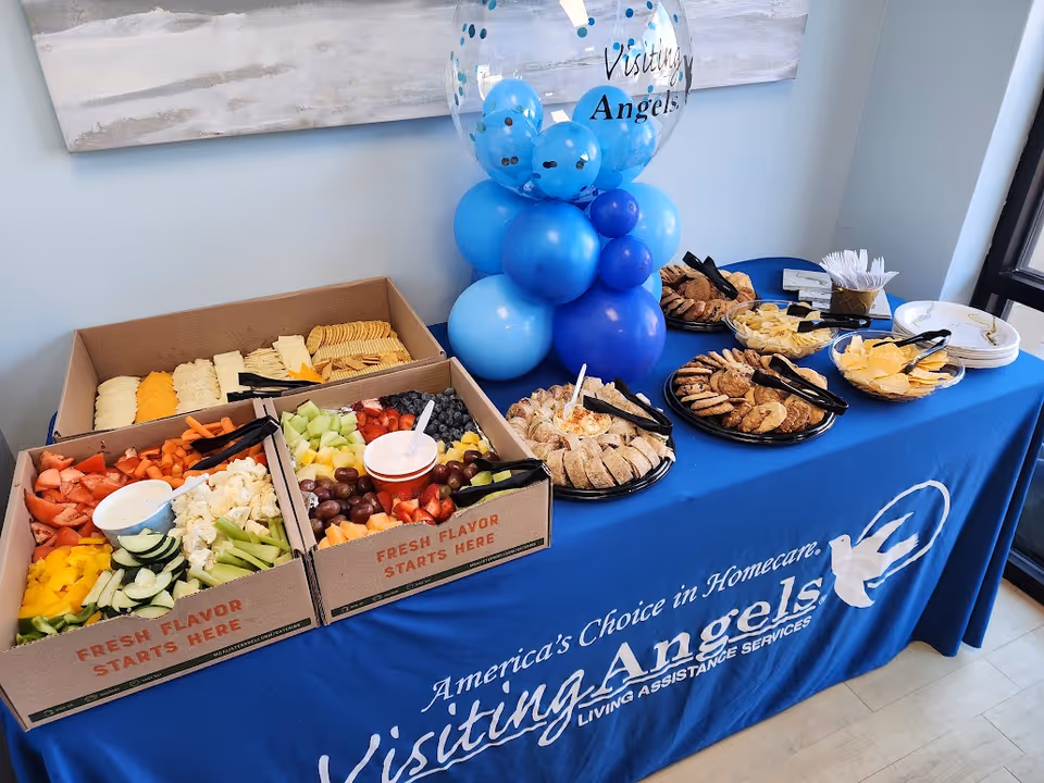 Buffet table with assorted fruits, snacks, pastries, and blue balloons at Visiting Angels.