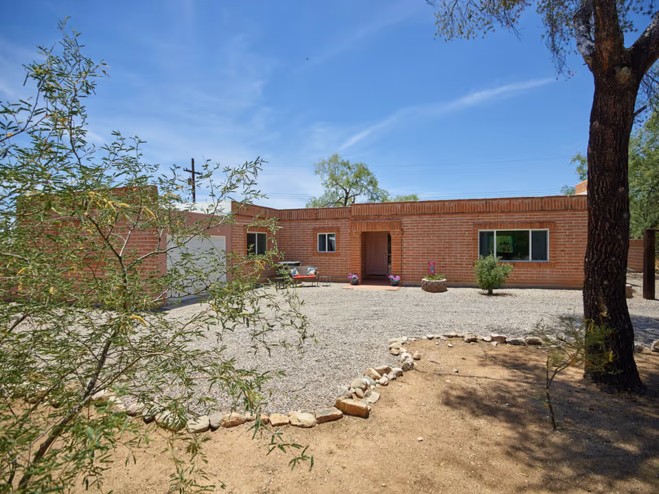 Single-story brick building with a gravel front courtyard, desert landscaping and a tree under a blue sky.