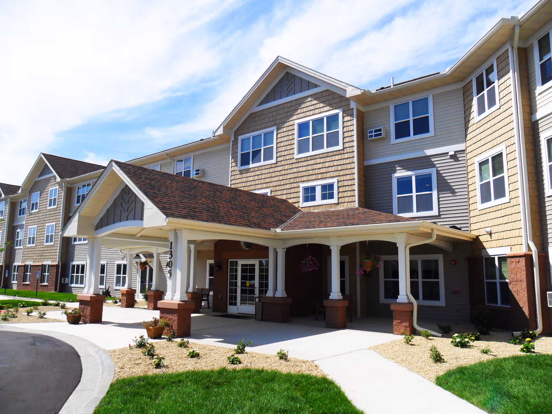 Exterior view of a multi-story senior living facility building with a covered entrance, multiple windows, and landscaped grounds under a partly cloudy sky.