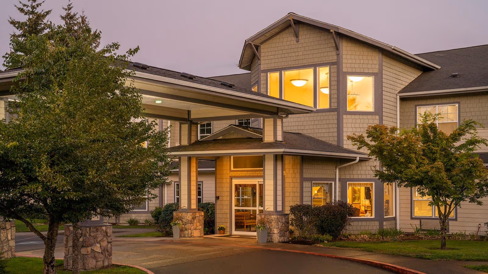 Front entrance of a two-story assisted living building with a covered drop-off canopy and warm interior lights at dusk.