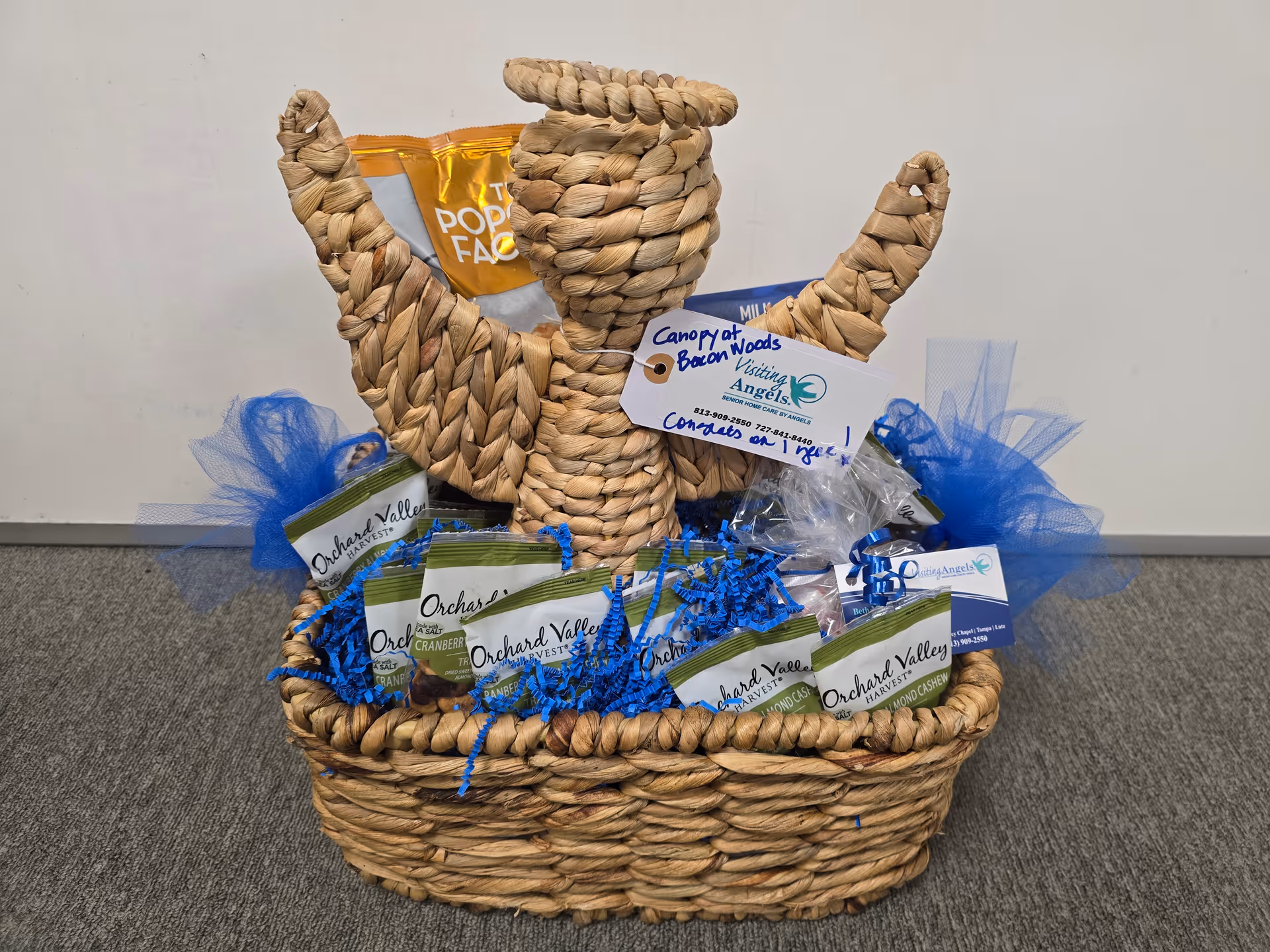 Woven gift basket with blue accents sits indoors, labeled Visiting Angels.