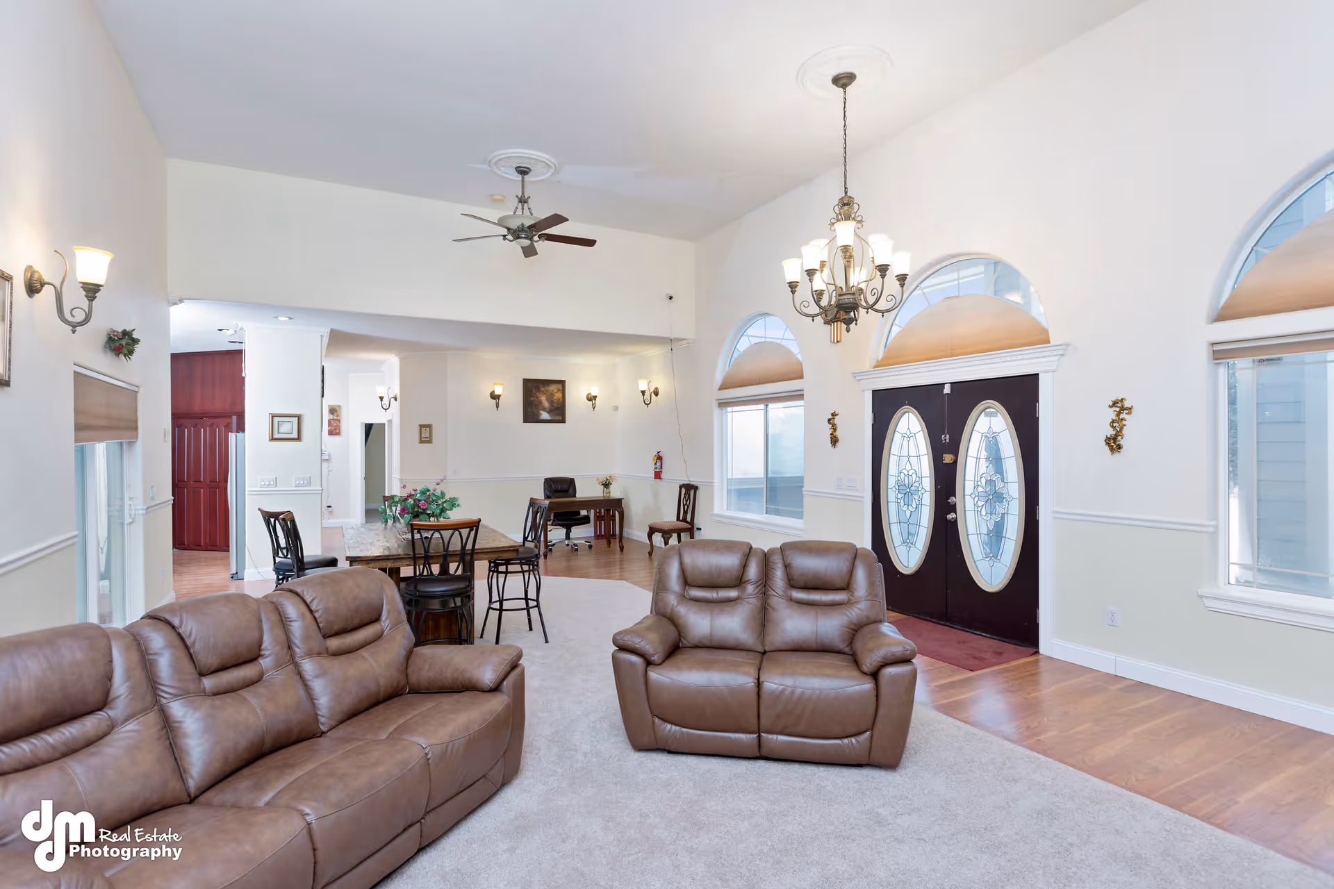 Spacious living room area with two brown leather sofas on a beige carpet. The room features high ceilings with a ceiling fan and a chandelier. There are large arched windows with blinds and a double front door with decorative glass panels. In the background, there is a dining table with chairs and a small desk with a chair against the wall. The walls are painted white with wall sconces providing additional lighting.