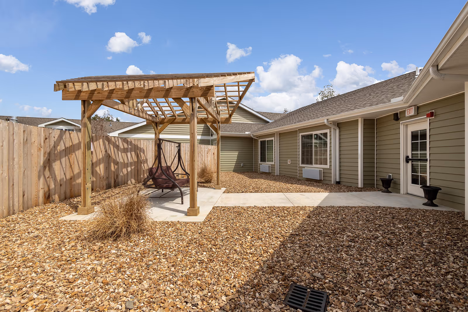 Outdoor courtyard area with a wooden pergola covering two brown swinging chairs. The ground is covered with small rocks and there are some dry plants near the pergola. The courtyard is enclosed by a wooden fence and the exterior walls of the building are visible, painted in a light green color with white trim. The sky is blue with some scattered clouds.