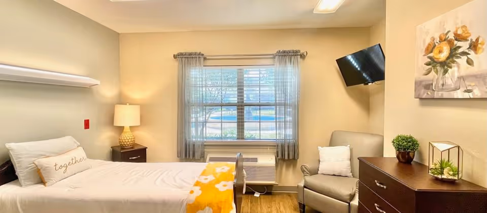 A bright and cozy bedroom with a single bed featuring white bedding and a yellow floral blanket. There is a pillow with the word 'together' on it. Next to the bed is a nightstand with a lamp. A window with sheer curtains lets in natural light. Across from the bed is a comfortable armchair with a white pillow, a dresser with decorative plants, and a wall-mounted flat-screen TV. A floral painting hangs on the wall above the dresser.
