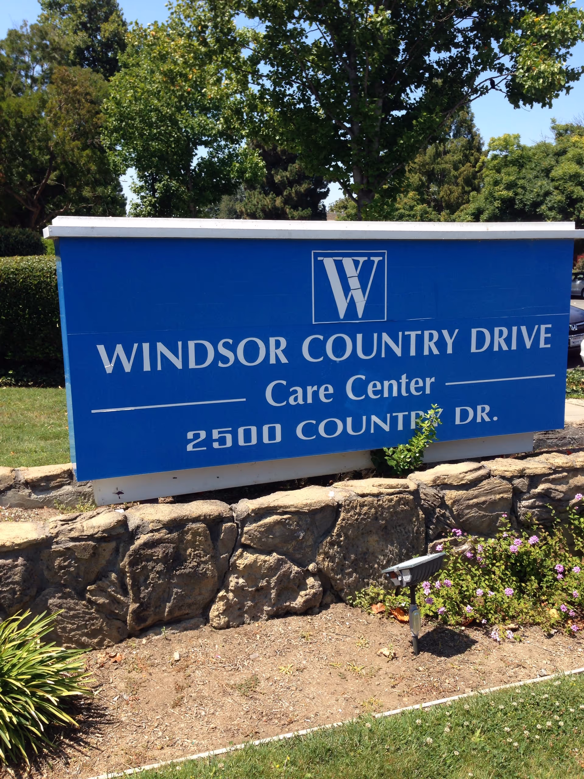 Blue Windsor Country Drive Care Center sign on a stone wall with plants.
