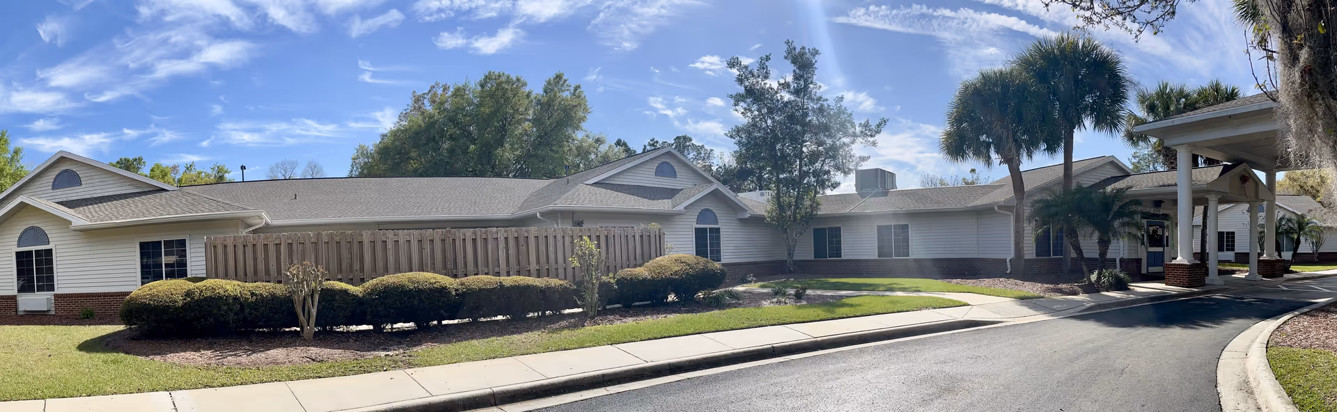 Single-story senior living building with a covered entrance, landscaped shrubs and palm trees under a blue sky.